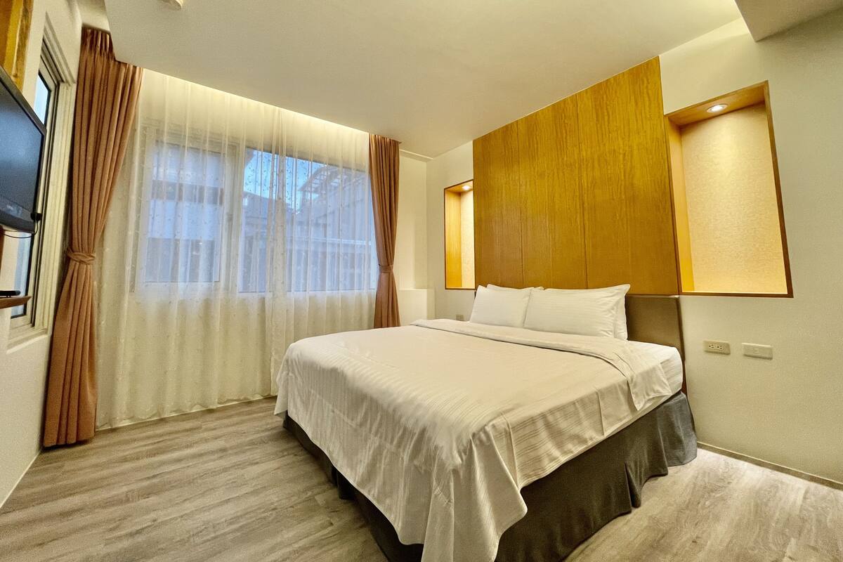 Habitación de hotel con una cama grande, cabecero de madera, televisor y ventana con cortinas.