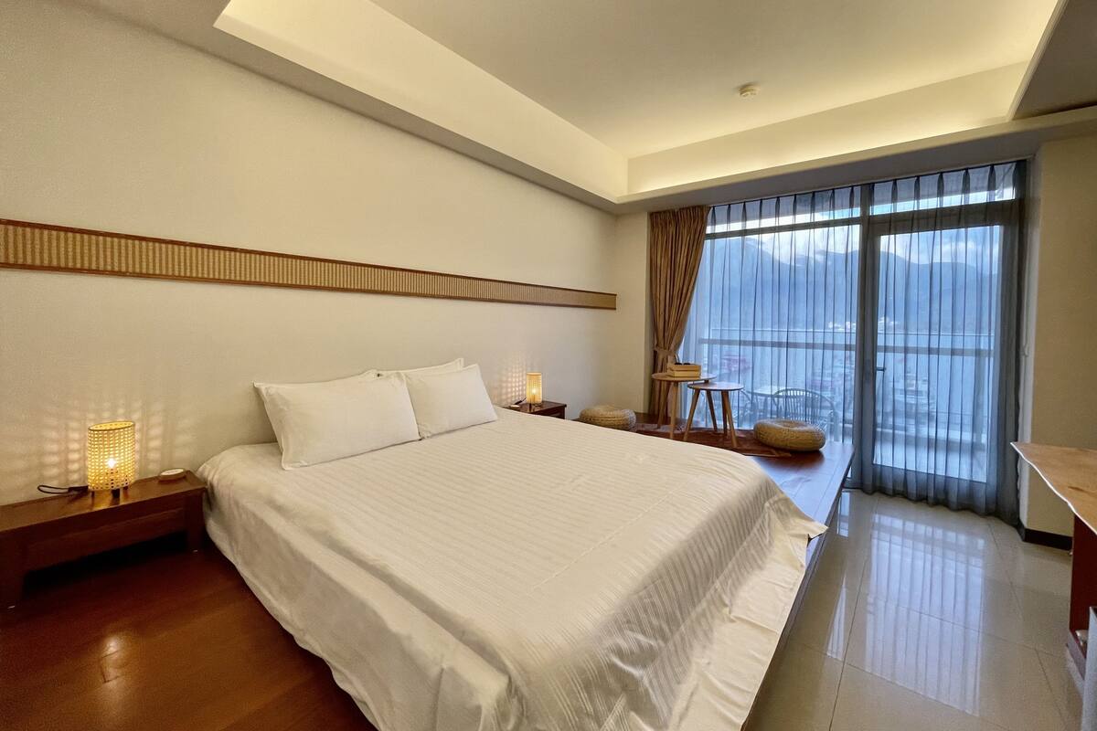Una habitación de hotel moderna con una cama grande, una mesita de noche, una pequeña mesa con lámpara y vistas al exterior a través de un amplio ventanal.
