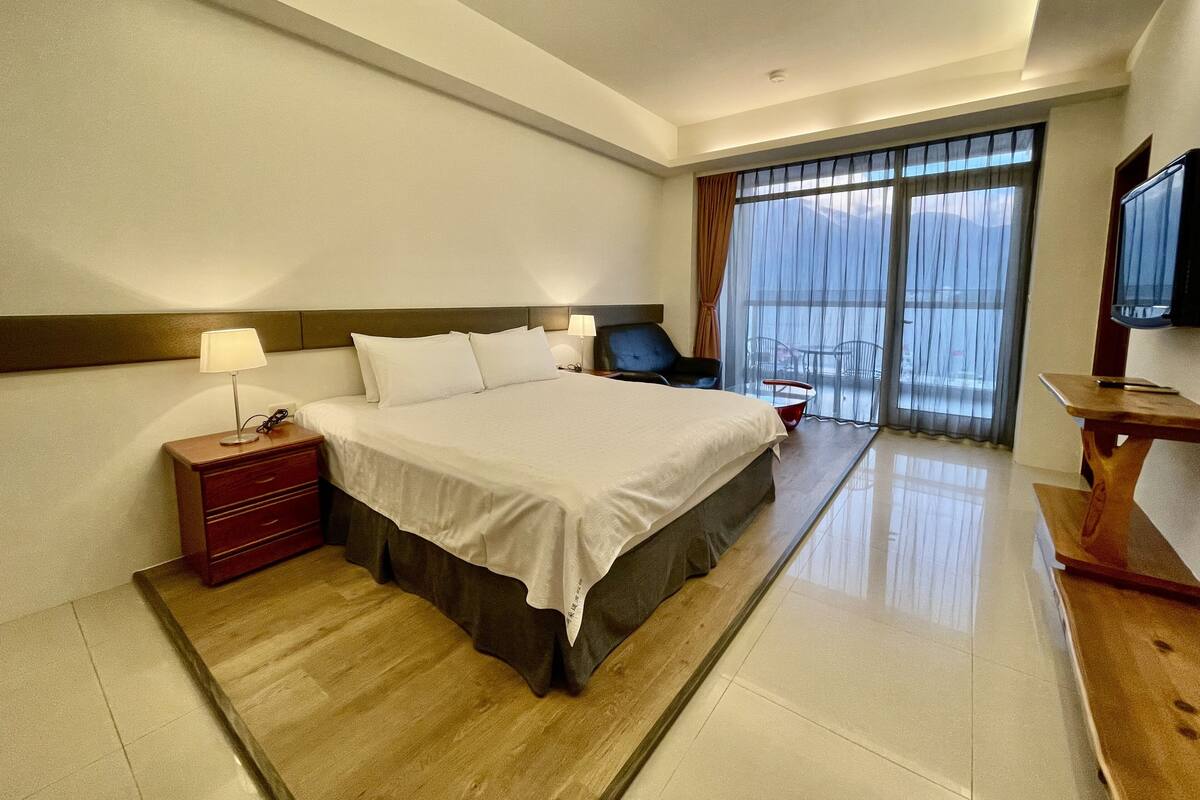 Habitación de hotel con una cama grande, mesitas de noche, un televisor y un balcón con vistas.