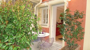 A small outdoor seating area with a table and chairs, surrounded by greenery and a door leading to an interior space.