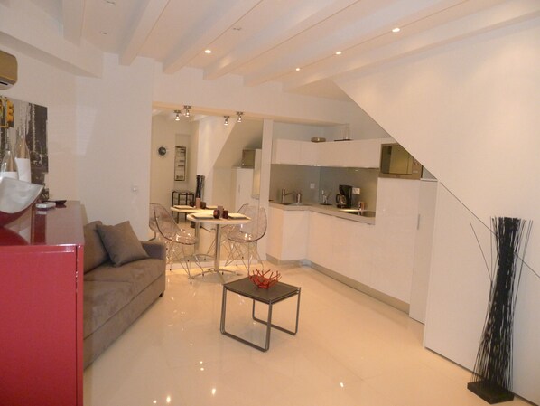 A modern living room with a sofa, a coffee table, and a dining area with chairs and a table. There is a kitchen area with cabinets and a microwave.