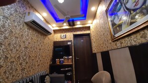 A room with patterned wallpaper, a ceiling fan, recessed lighting, an air conditioning unit, a door with a number, and a framed picture on the wall. - Krishna Inn (New Delhi)