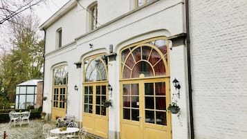 Un bâtiment blanc doté de grandes fenêtres en arc de cercle et d’une porte jaune. On y trouve des tables et des chaises à l’extérieur.
