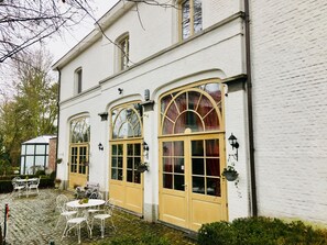 A white building with large arched windows and a yellow door. There are outdoor tables and chairs. - Koetshuis Bosdam B&B (Beveren)