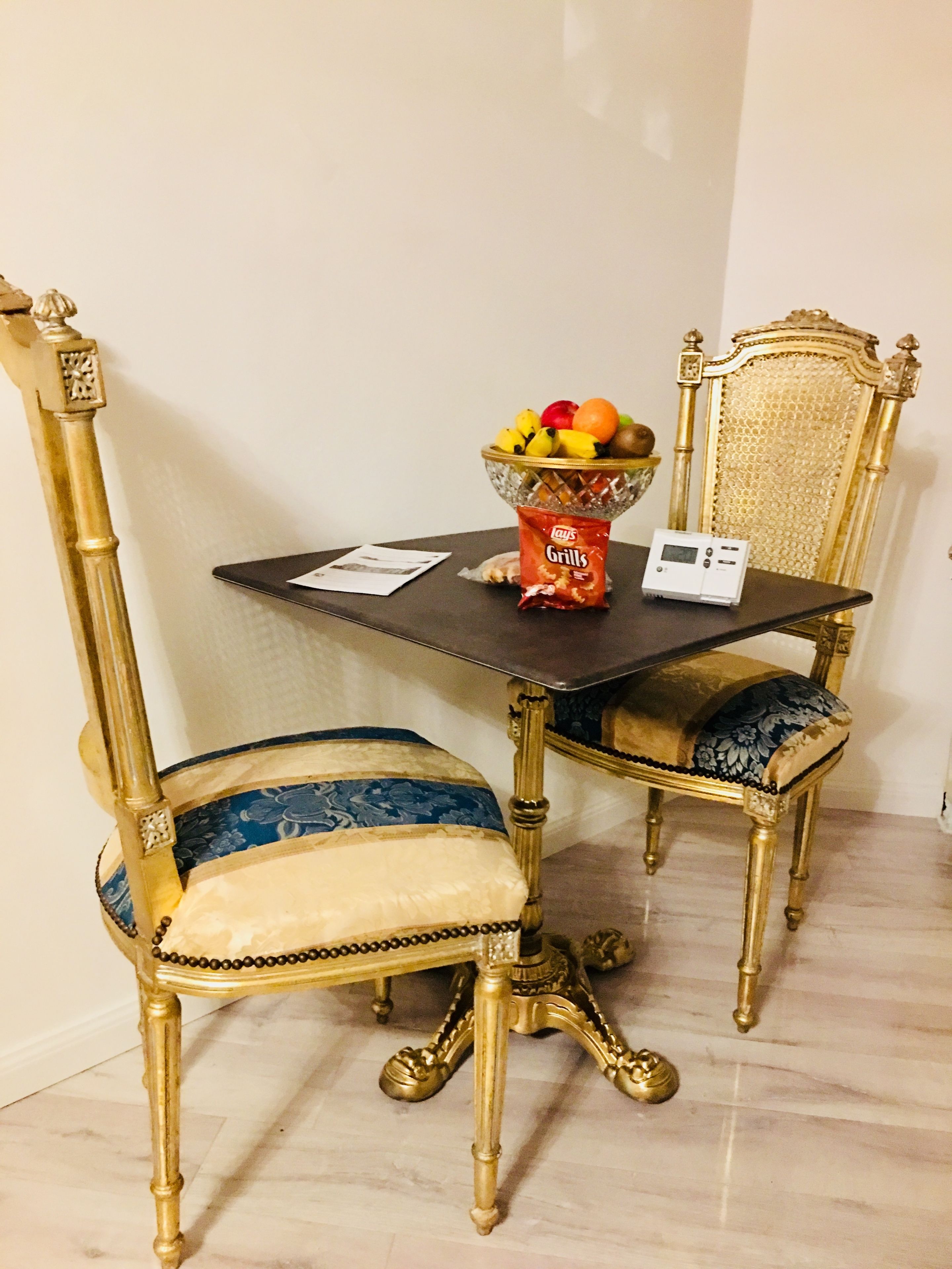Una mesa de comedor con un cuenco de fruta y un paquete de papas fritas. Hay dos sillas decorativas con tapicería azul y dorada.