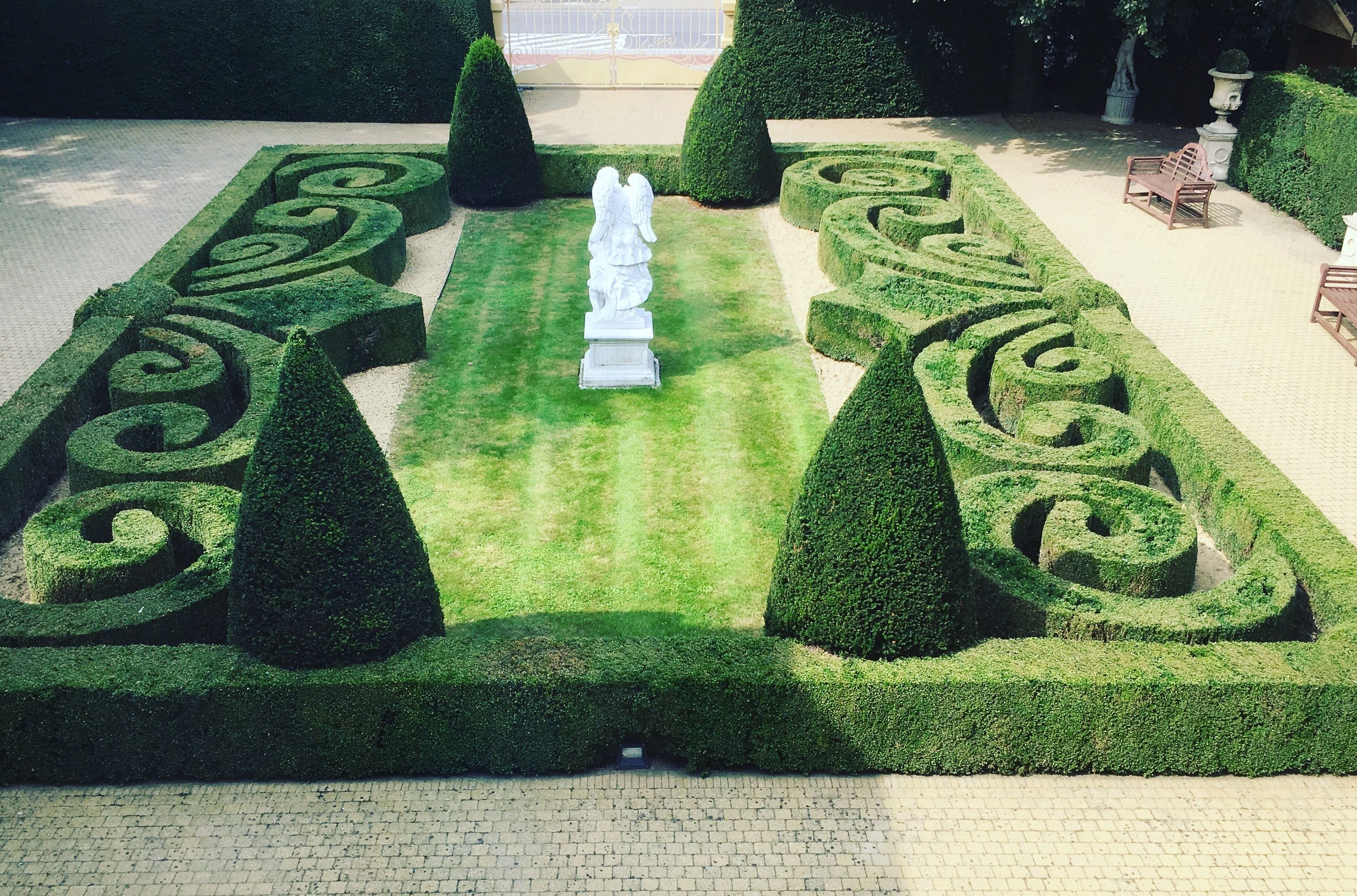Un jardín meticulosamente cuidado con patrones geométricos de setos, una estatua central y zonas de estar.