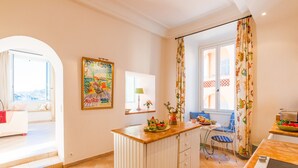 Una cocina luminosa con una isla central, un comedor con una silla azul y vistas a través de una ventana.
