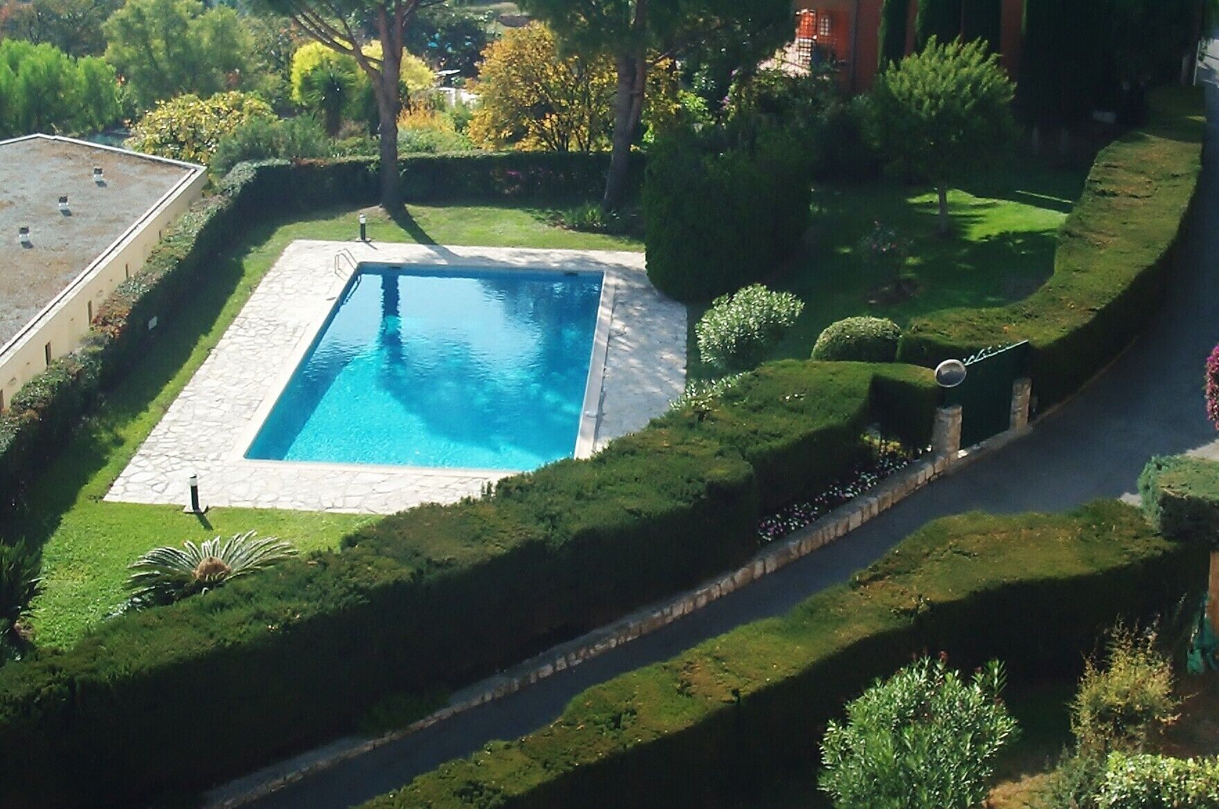 A rectangular pool with a stone border, surrounded by well-manicured grass and hedges, with a paved walkway and a small garden area.