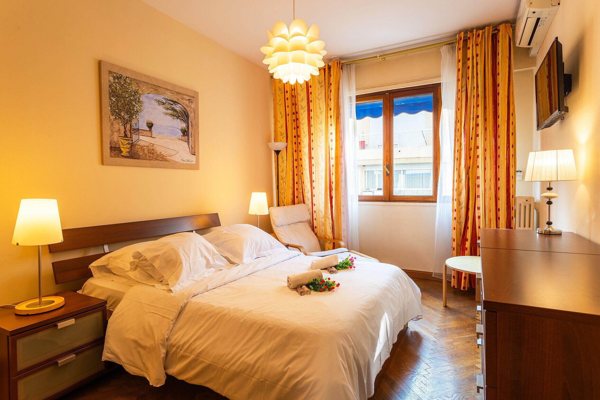 A hotel room with a bed, bedside tables, a desk, a chair, a window with curtains, and a painting on the wall.