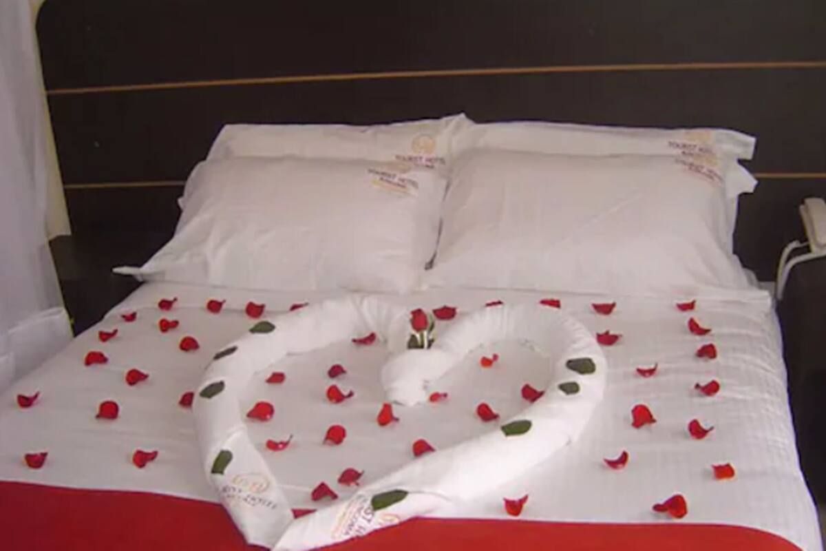 A neatly made bed with white linens and red rose petals arranged in a heart shape.