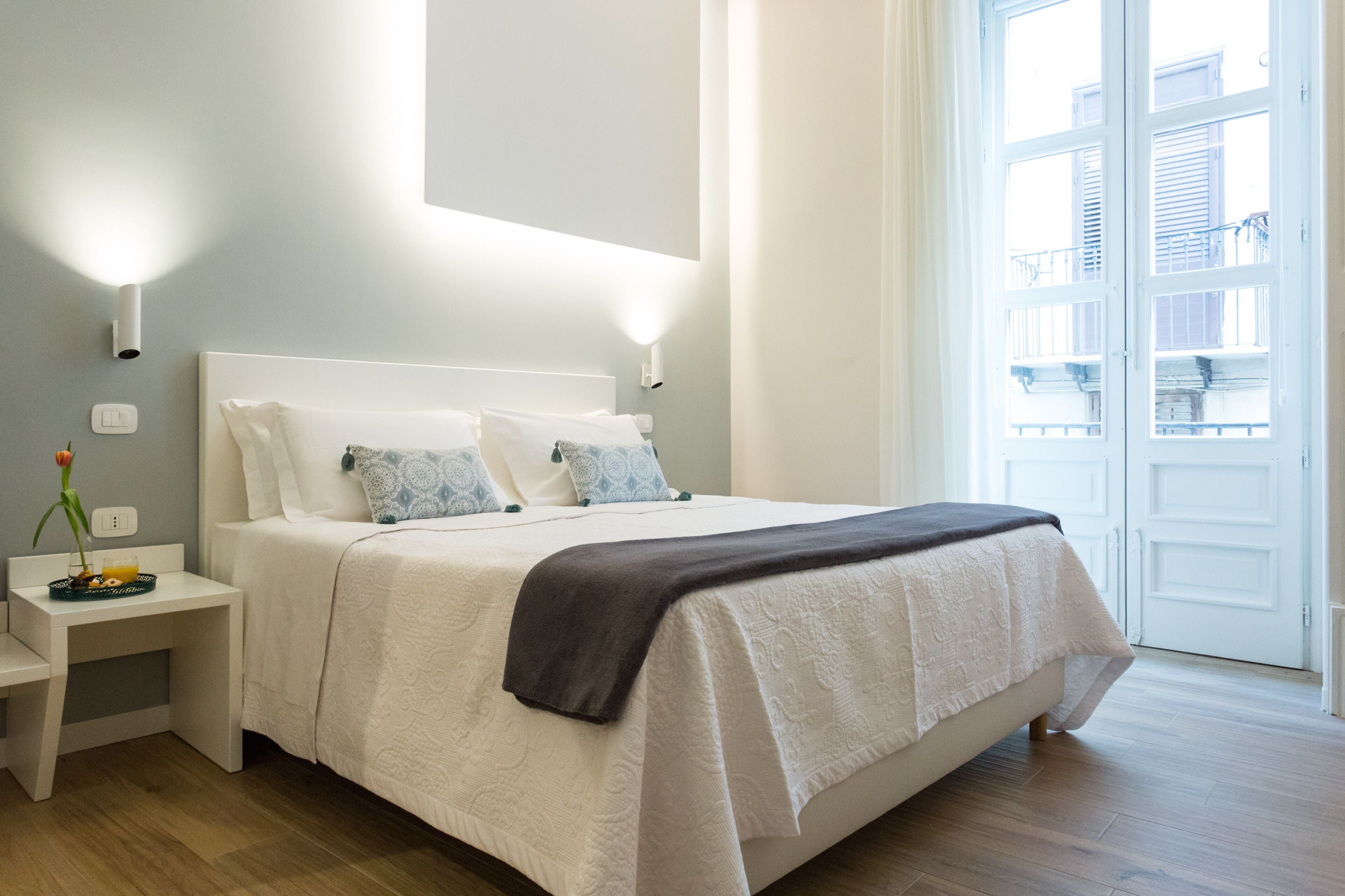 Una cama bien hecha con cabecera blanca, una mesita de noche con un florero y un frutero, y un ventanal con vistas a edificios.