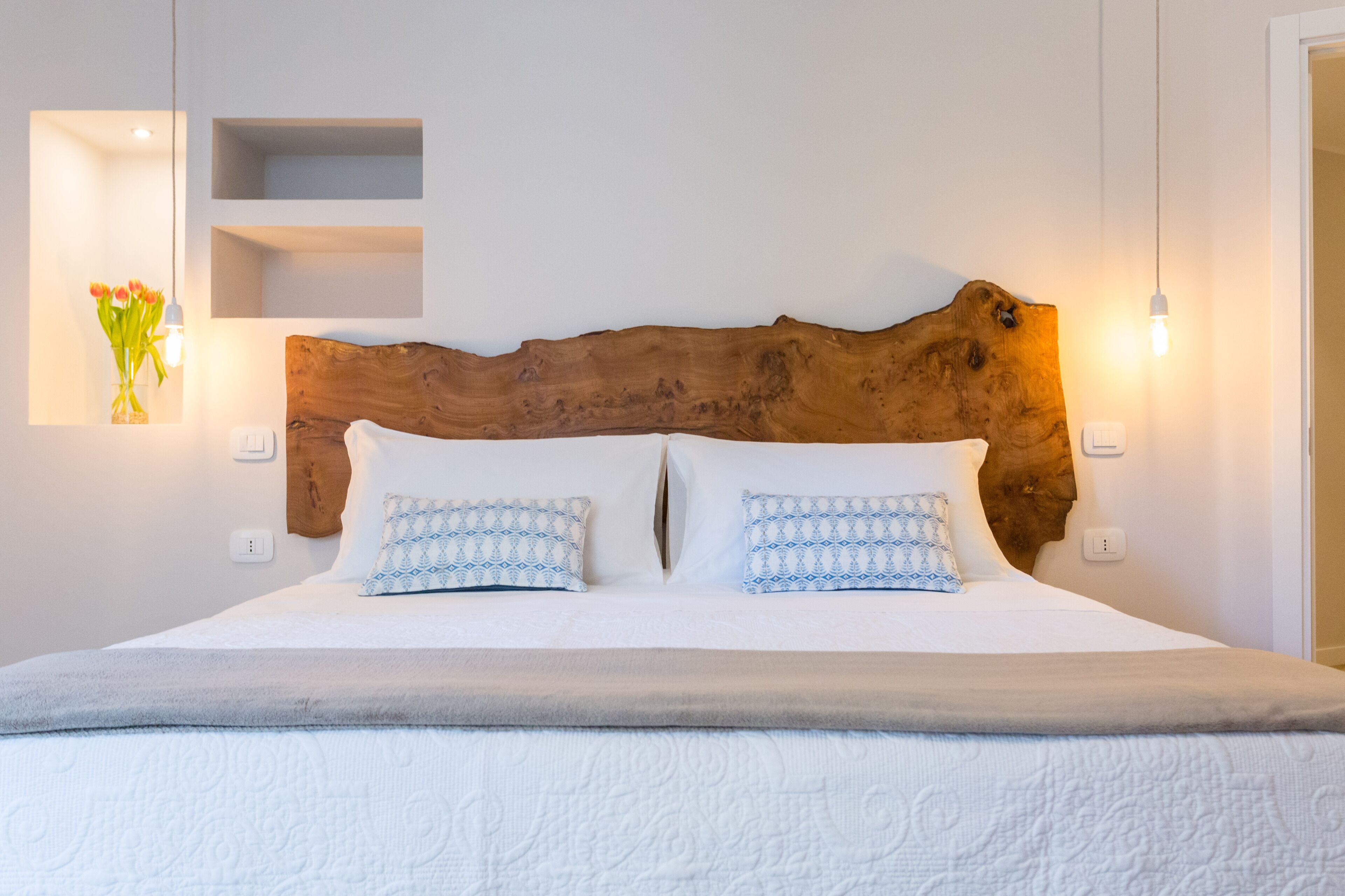 A bedroom with a wooden headboard, two patterned pillows, a white bedspread, and a vase with flowers on the left wall.