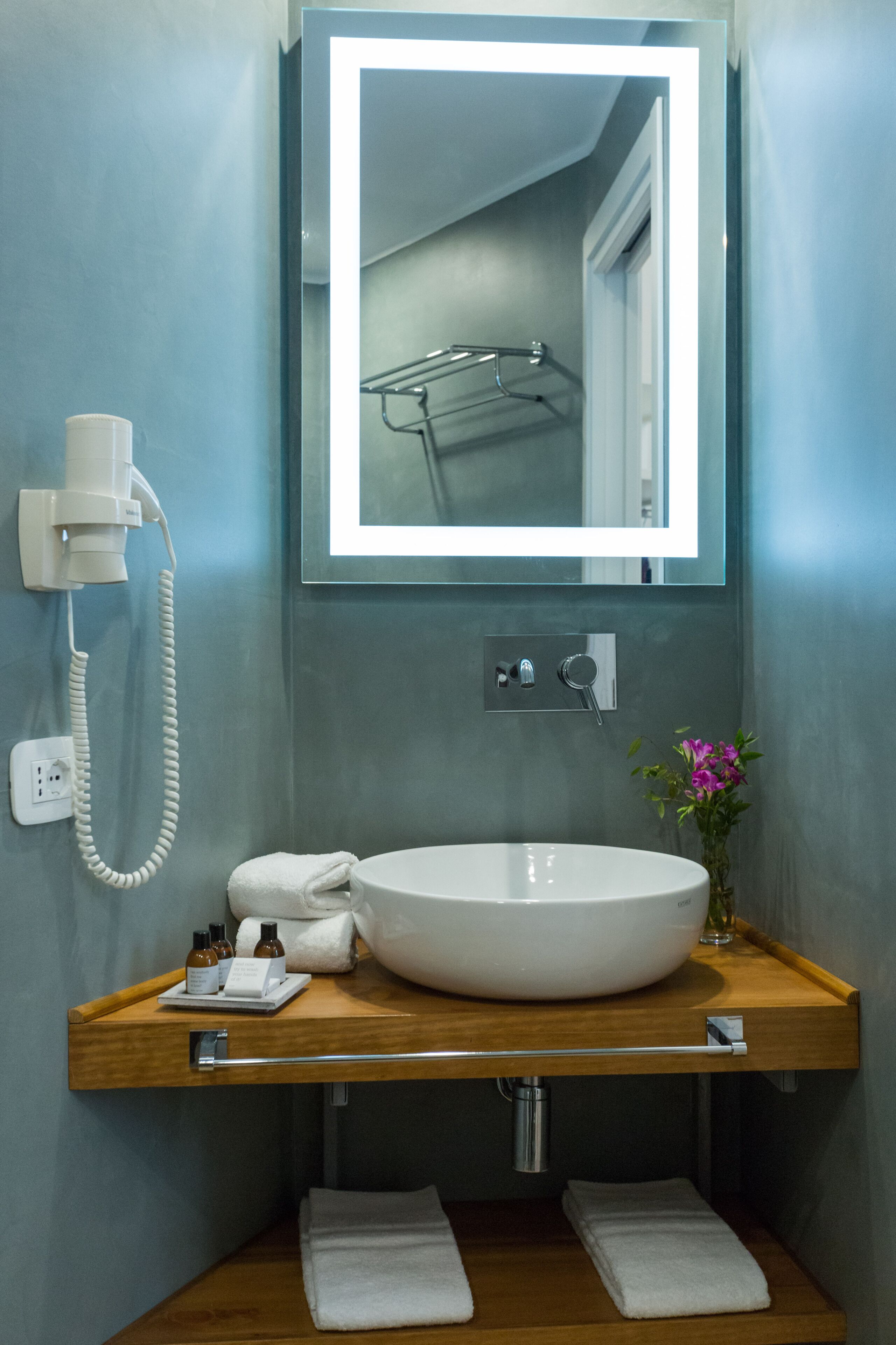 A bathroom with a round white sink, a mirror, a towel rack, a hairdryer, and a small vase with flowers.