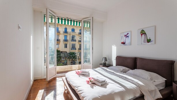 Quarto com uma cama grande, uma mesinha de cabeceira, uma luminária e vista para um edifício através de uma porta de vidro.