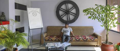Un hombre sentado en un sofá en una habitación con un adorno grande de rueda, una estatua de Buda y una planta en maceta.