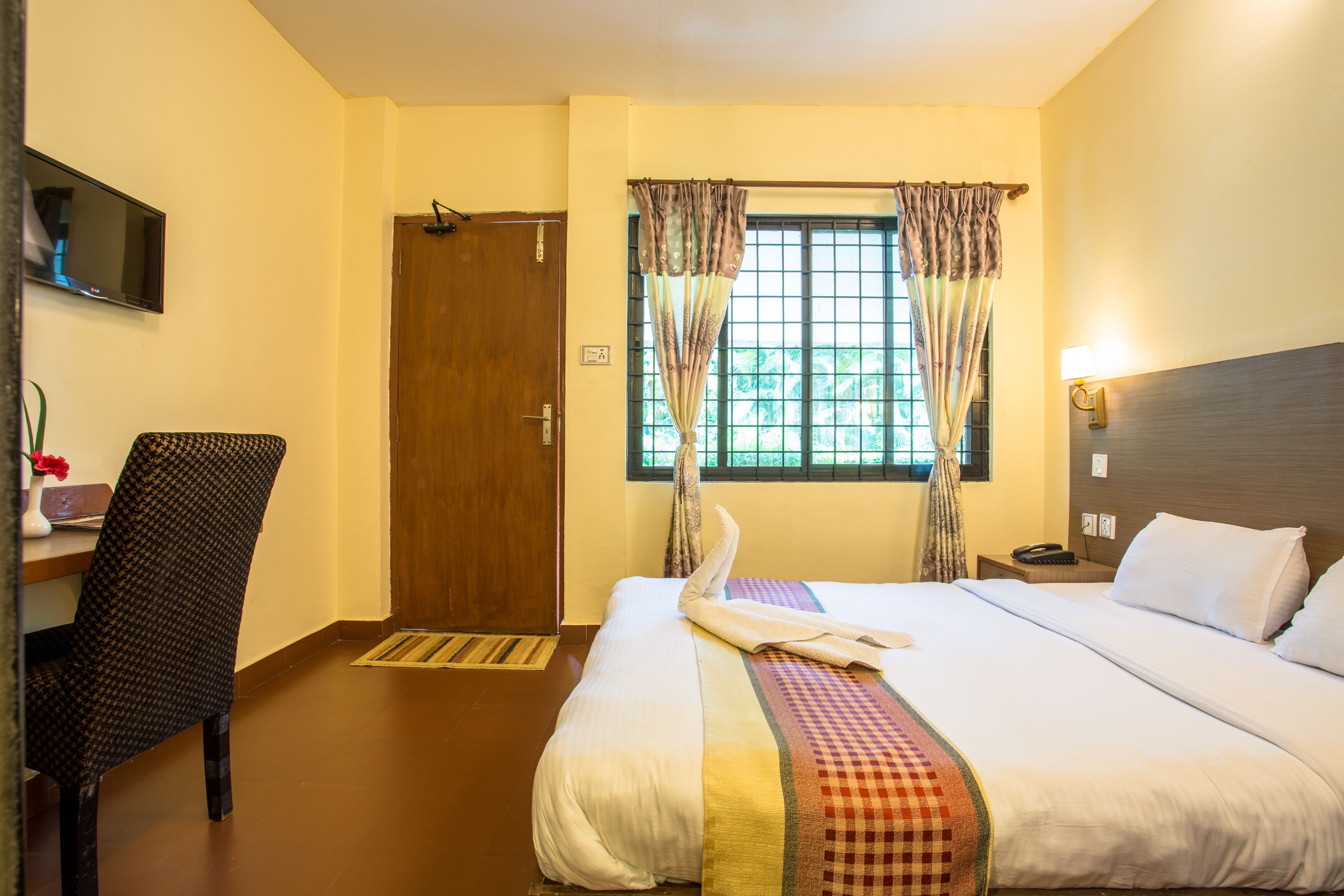 A hotel room with a bed, a chair, a desk with a lamp, a TV, and a window with curtains.