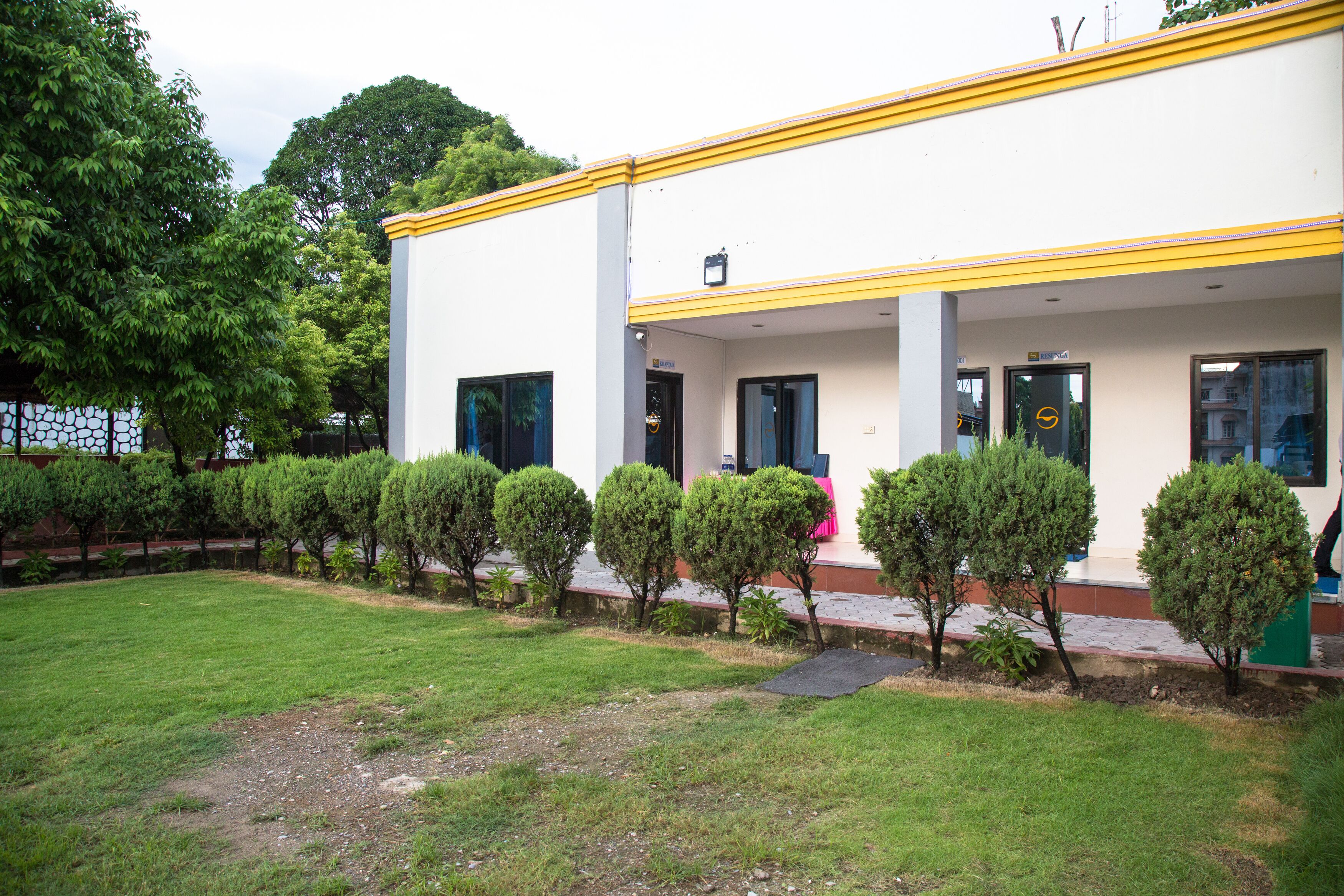 Um edifício com fachada branca e detalhes em amarelo, entrada coberta e um gramado bem cuidado, com arbustos aparados.