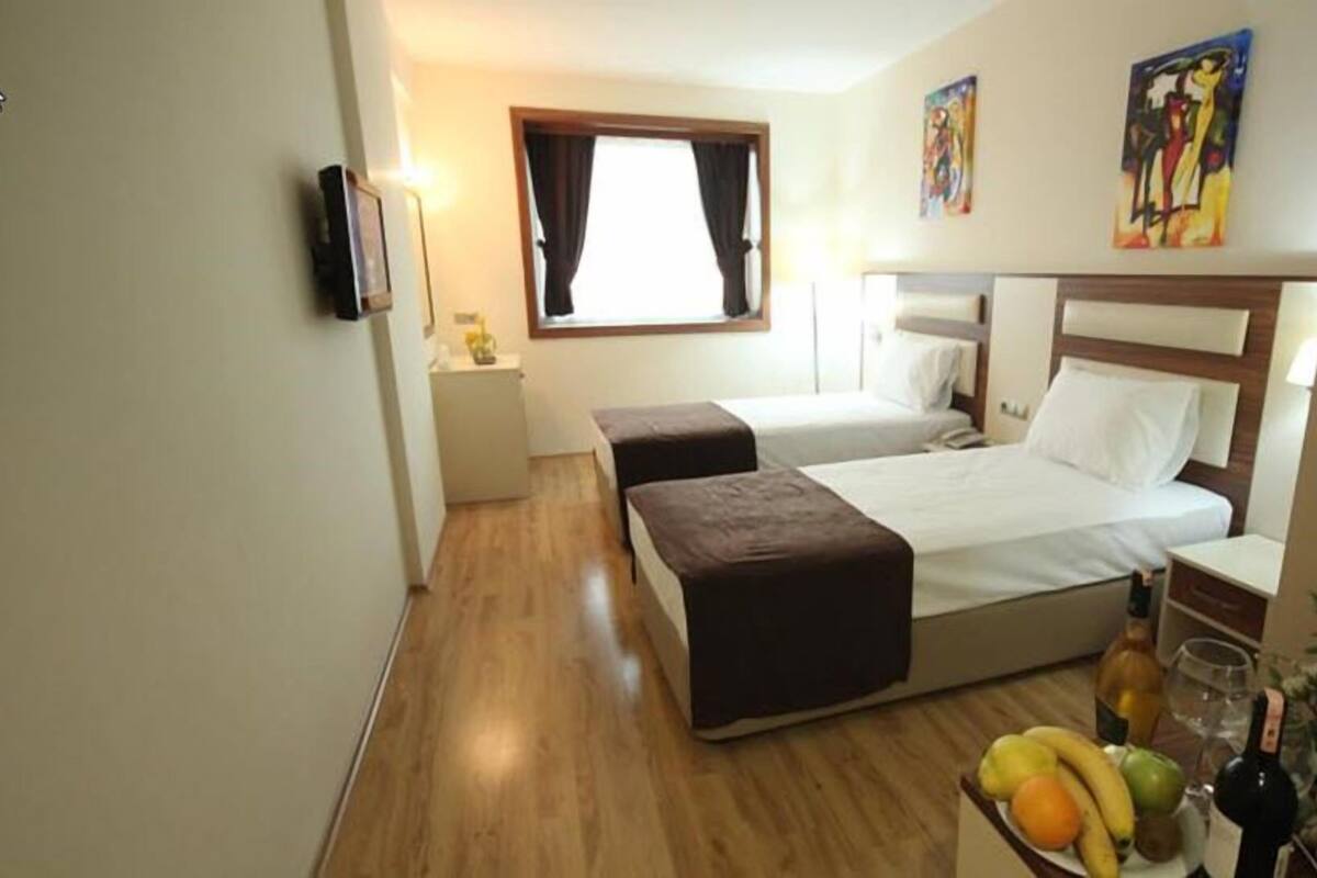 Une chambre d’hôtel avec deux lits, une petite table avec des fruits et une bouteille, une table de chevet, une télévision et des œuvres d’art accrochées aux murs.