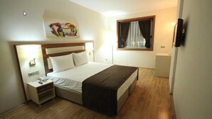 A hotel room with a bed, bedside tables, a nightstand, a window with curtains, and a wall-mounted TV.