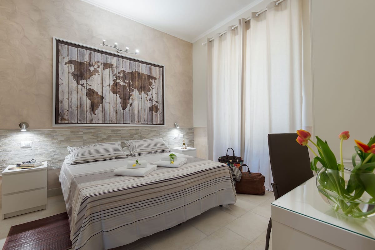 A modern bedroom with a large bed, bedside lamps, a world map artwork, a desk with a laptop, and a vase with flowers.
