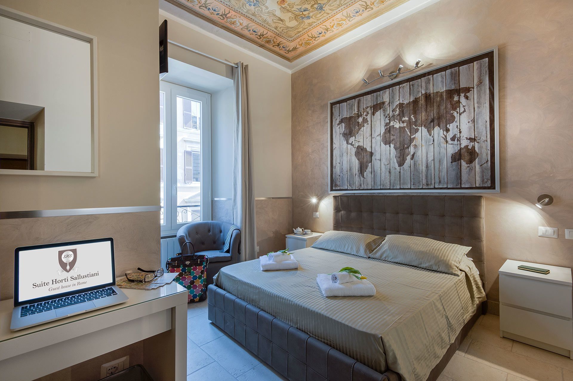 A modern bedroom with a large bed, a desk with a laptop, a world map artwork, and a window with a view of buildings.