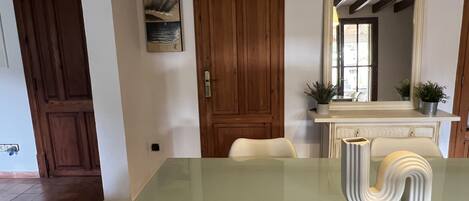 A dining area with a glass table, wooden doors, a painting, a mirror, and a fireplace with plants.