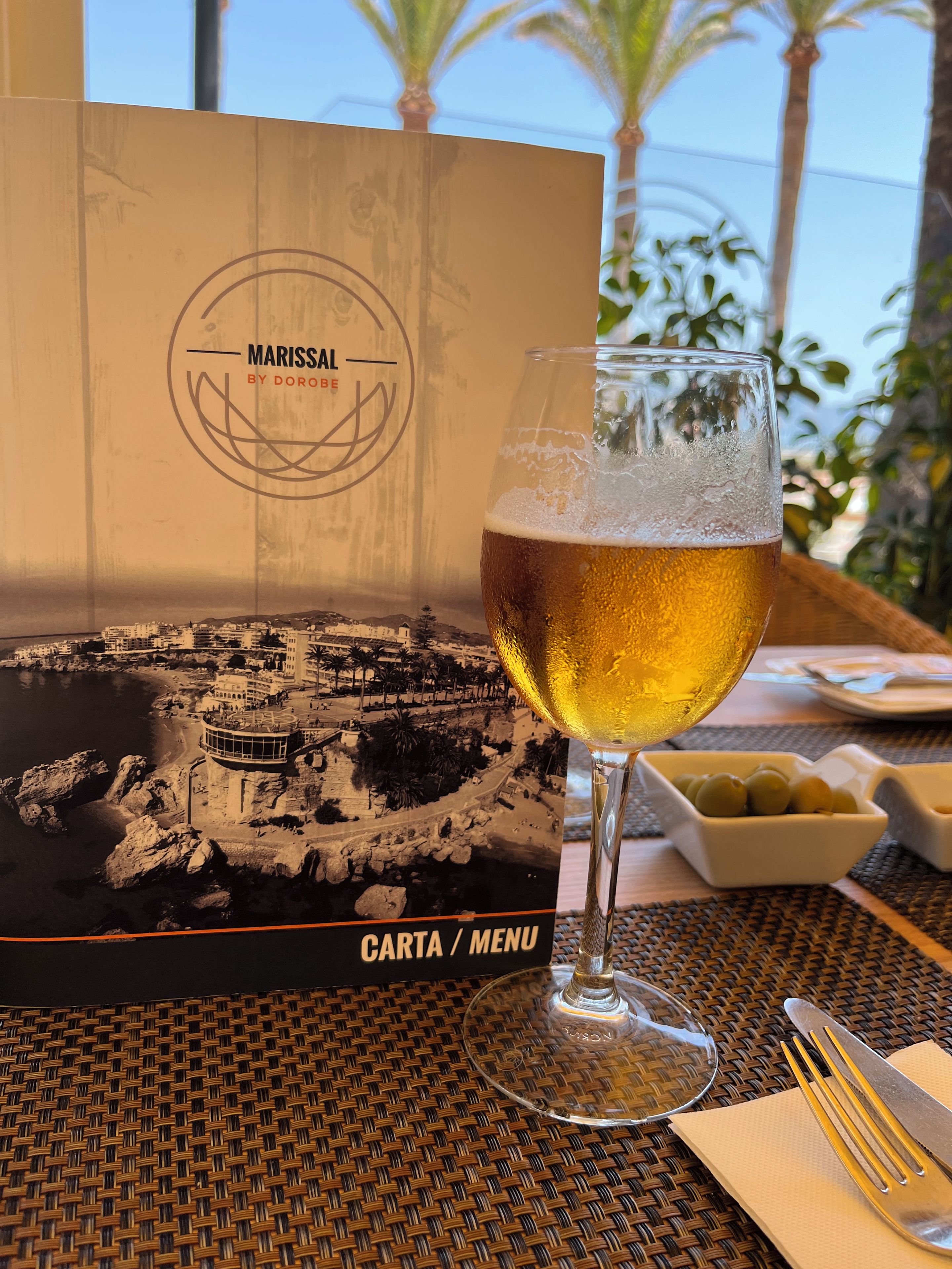 Um copo de cerveja sobre uma mesa com um menu e um postal mostrando uma cidade costeira.