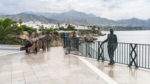 A coastal promenade with a statue, a cannon, and a view of a town and mountains. - Marissal (Nerja)