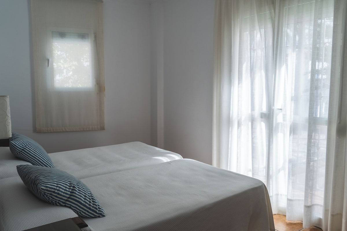 Quarto com cama, duas almofadas, janela e cortinas leves.