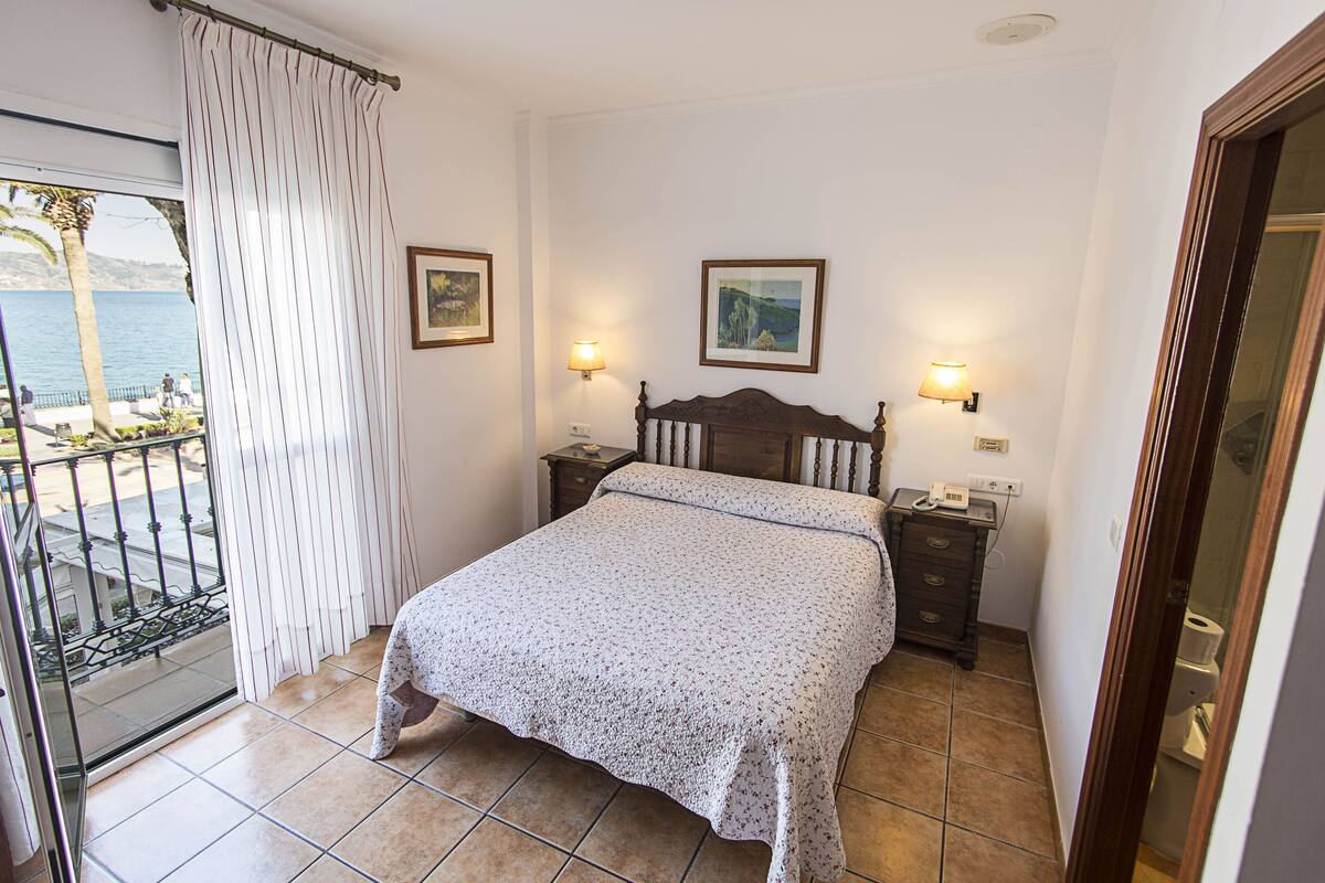 Quarto com cama, duas mesas de cabeceira, um criado-mudo, varanda com vista para o mar e duas luminárias de parede.