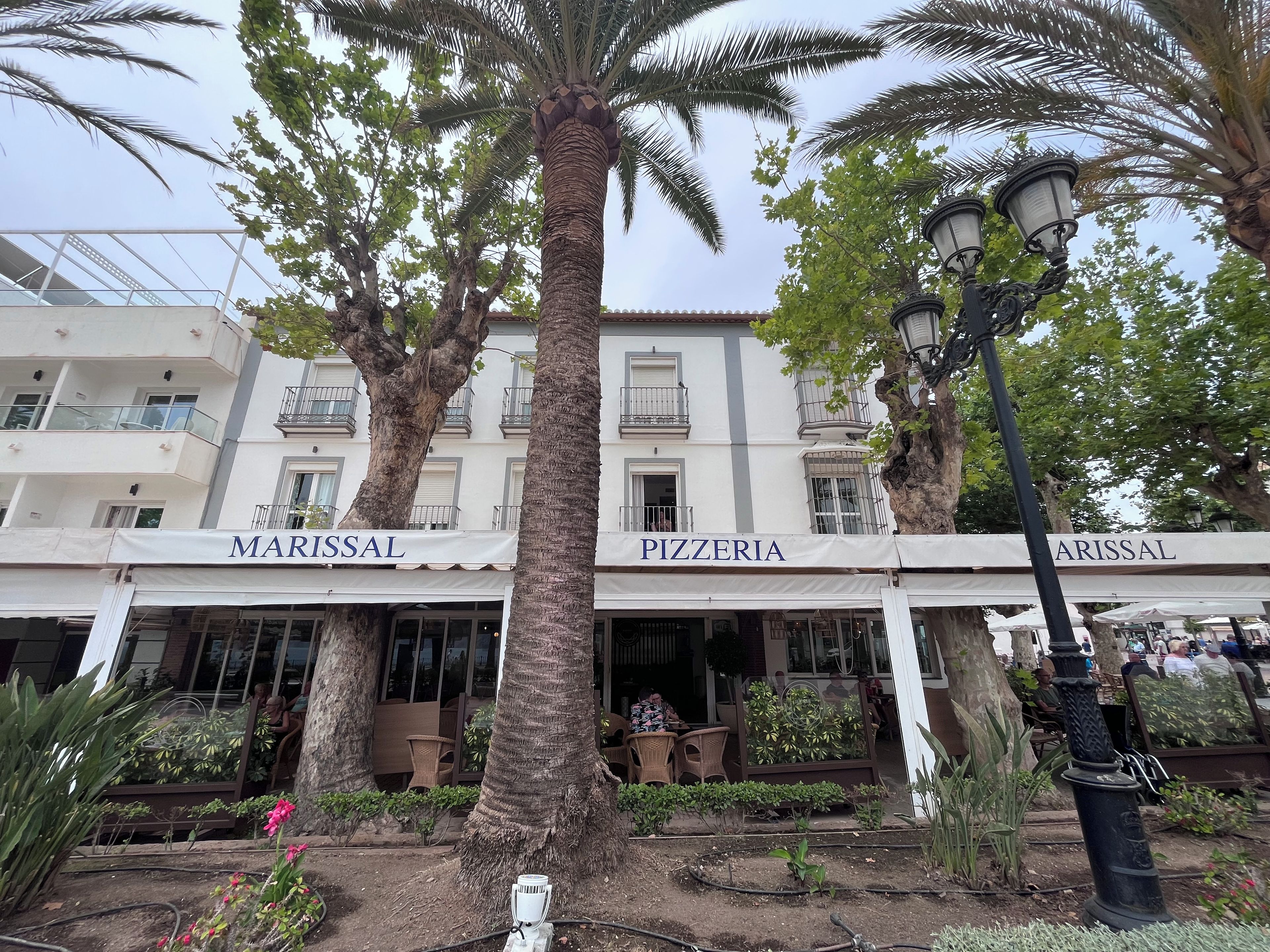 A building with 'MARISSAL' and 'PIZZERIA' signs, surrounded by palm trees and potted plants.