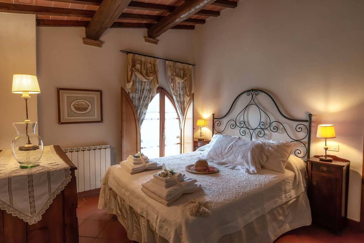 Ein Schlafzimmer mit einem Bett, zwei Nachttischlampen, einem Heizkörper, einem gerahmten Bild und einem Fenster mit Vorhängen.