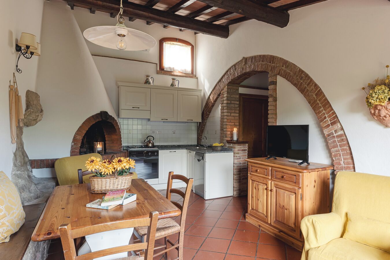 A cozy kitchen with a brick archway, a dining table with chairs, and a TV on a wooden cabinet.