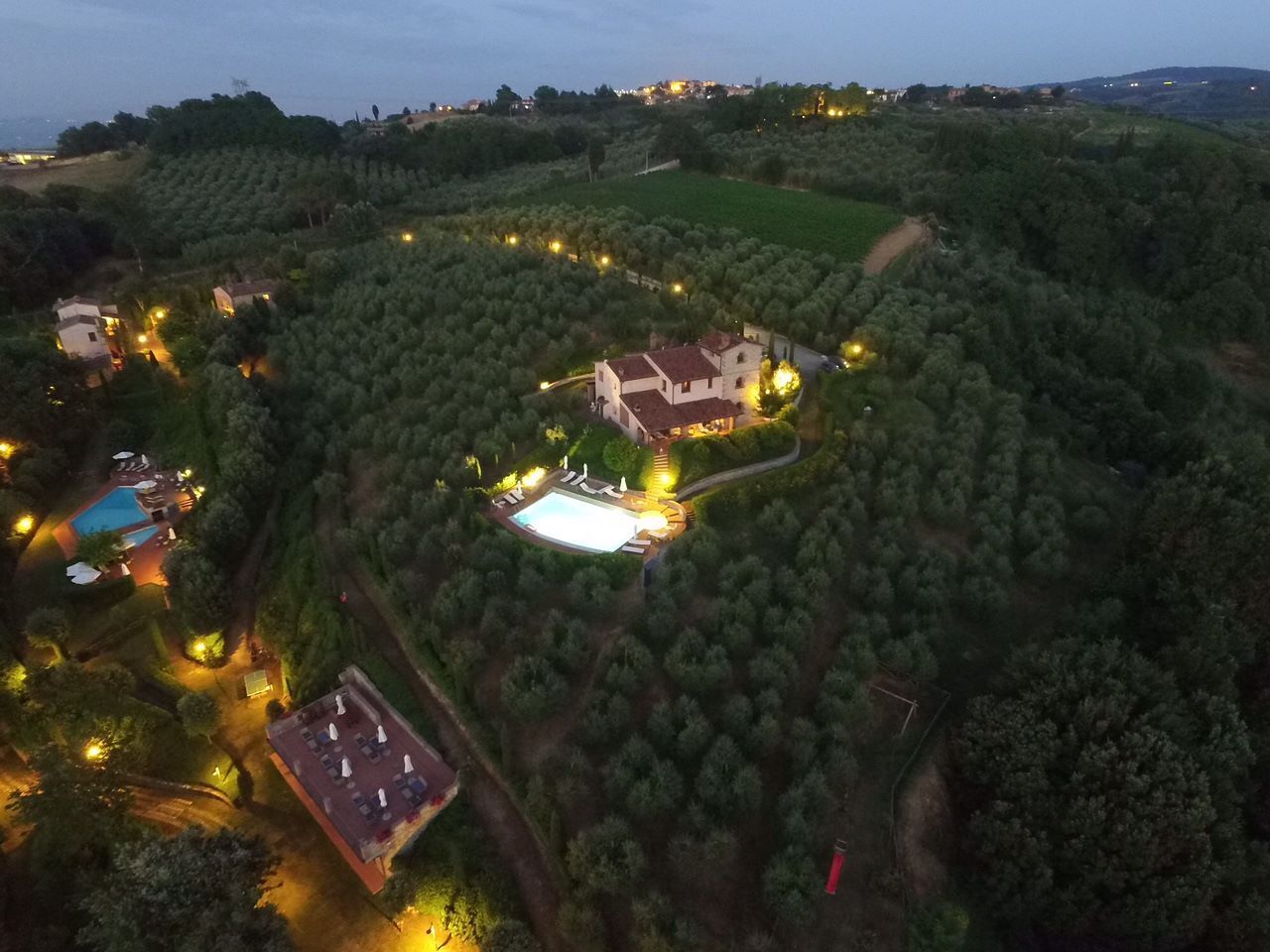 An aerial view of a property with a large house surrounded by trees, a swimming pool, and a tennis court.