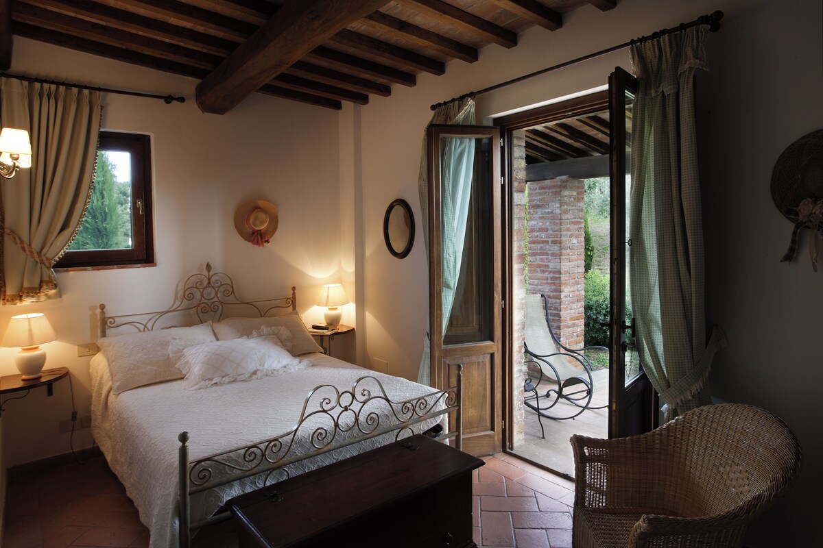 Une chambre à coucher avec un lit, des lampes de chevet, un fauteuil en osier et une vue sur l’extérieur, visible à travers une porte vitrée.