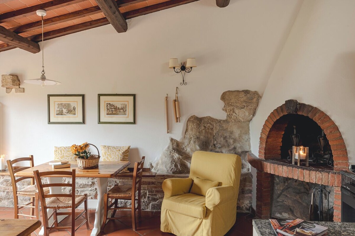 Un intérieur rustique avec une table à manger en bois, des chaises, un fauteuil jaune, un mur en pierre et une cheminée en briques.