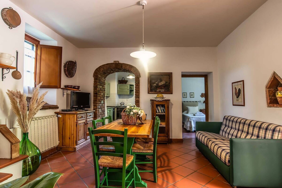 Un salon chaleureux avec un canapé vert, une table à manger en bois avec des chaises, un coin cuisine agrémenté d’une arche en briques et un luminaire suspendu.