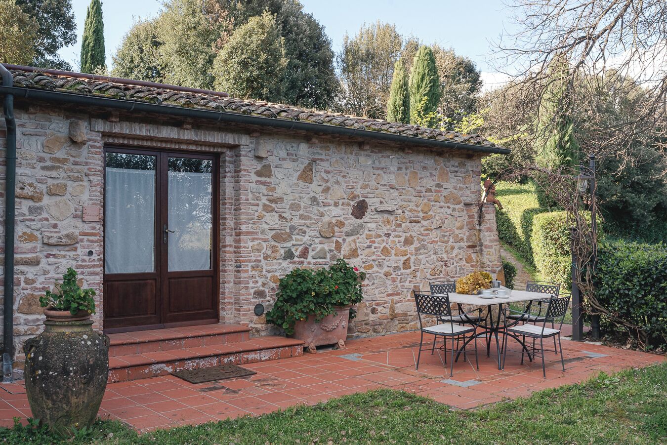 Un bâtiment en pierre avec un toit en tuiles, une porte en bois et un petit patio comprenant une table et des chaises.