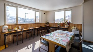 A dining area with wooden tables and chairs, large windows with a view of a snowy landscape, and a refrigerator in the corner. - Lodge Romulus (Rusutsu)
