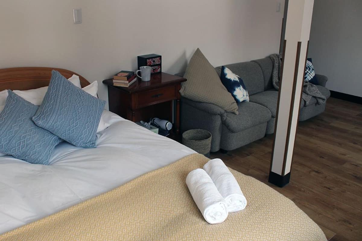 Uma cama bem arrumada com um manta bege e toalhas dobradas, um sofá cinza com almofadas estampadas, uma pequena mesa de cabeceira de madeira com livros e uma caneca, e piso de madeira.