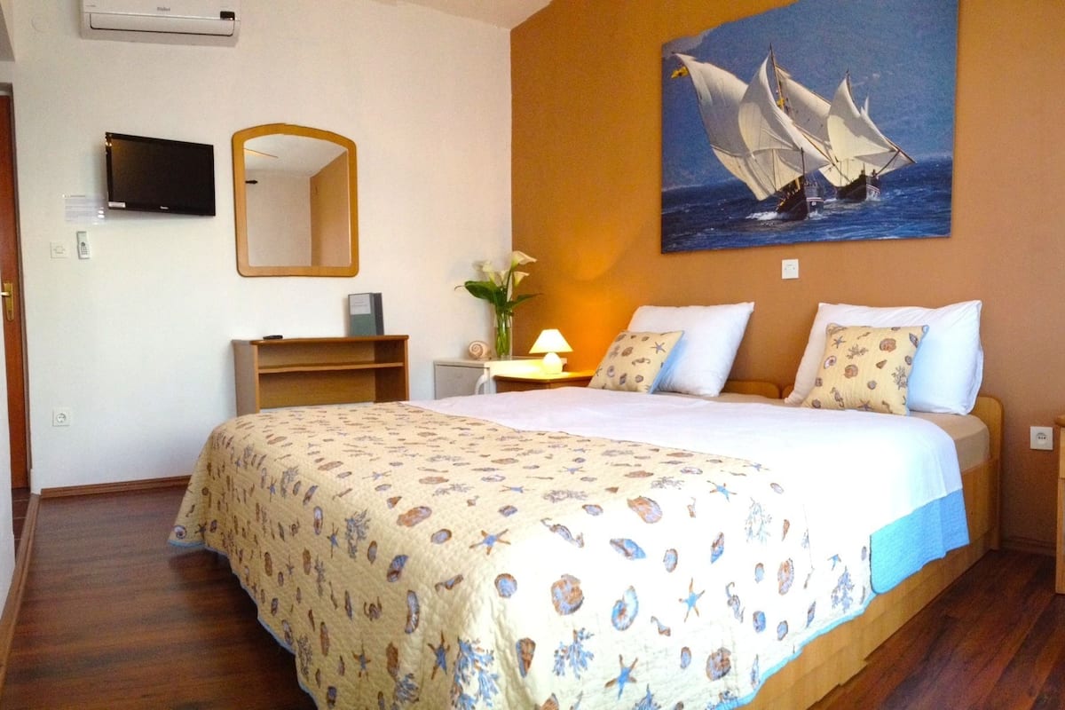 A hotel room with a bed, a desk, a chair, a mirror, a painting of sailboats, and a vase with flowers.