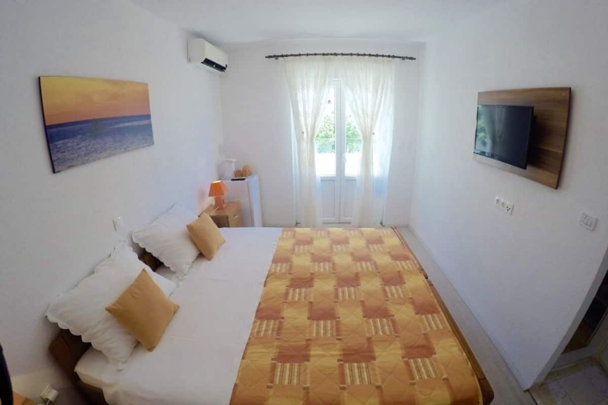 A bedroom with a bed, a TV mounted on the wall, a painting, and a window with curtains.