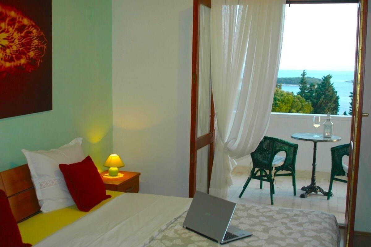 A hotel room with a bed, a laptop, a nightstand with a lamp, a window with curtains, and a balcony with chairs and a table.