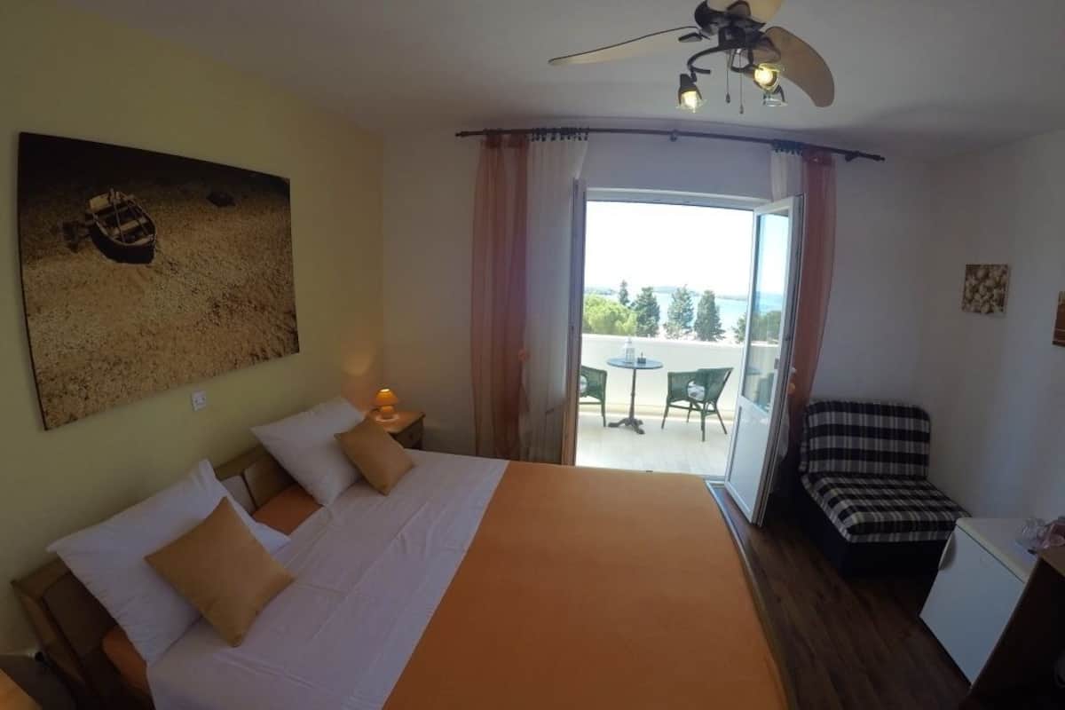 A hotel room with a bed, a ceiling fan, a small table with a lamp, and a view of the outdoors through a glass door.