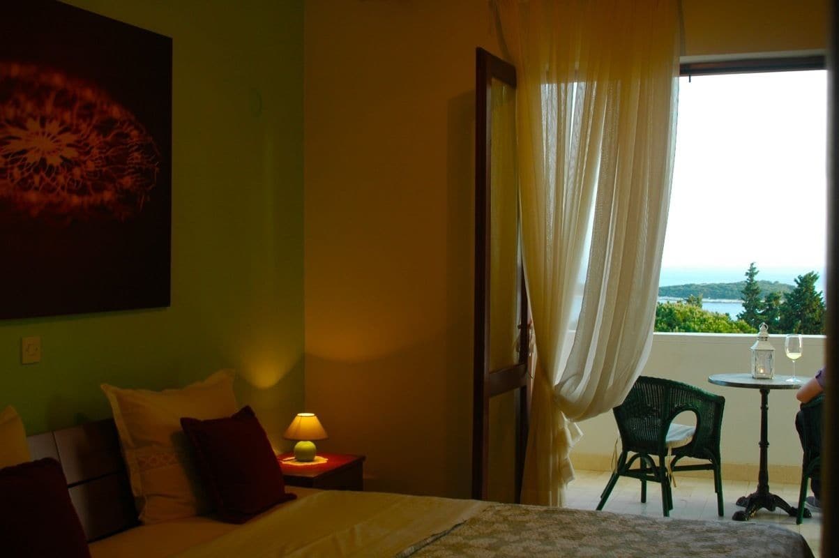A hotel room with a bed, a nightstand with a lamp, a chair, and a view of the sea through a window with curtains.