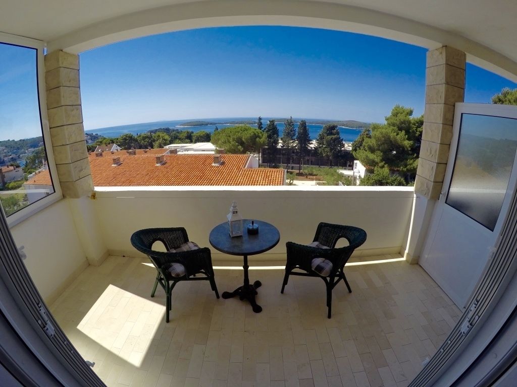 A balcony with two wicker chairs and a small round table, overlooking a coastal view with buildings and trees.