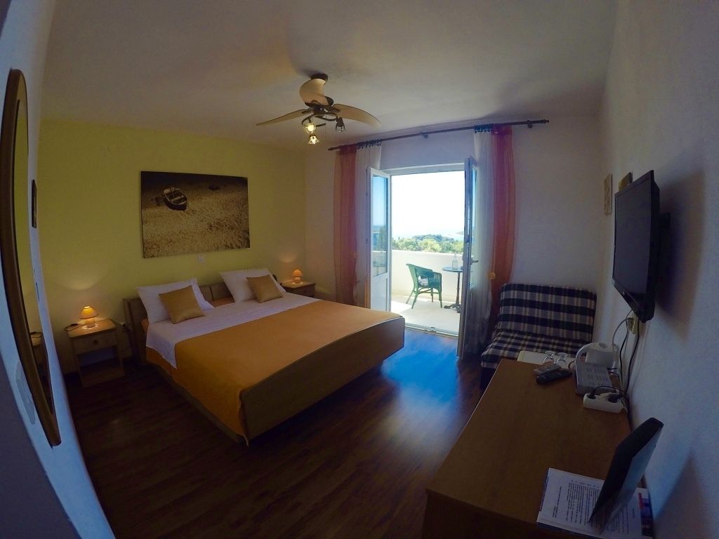 Quarto de hotel com cama, escrivaninha com computador, televisão, ventoinha no teto e janela com cortinas.
