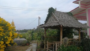 A traditional thatched-roof structure with a wooden fence, surrounded by potted plants and a yellow flowering bush.