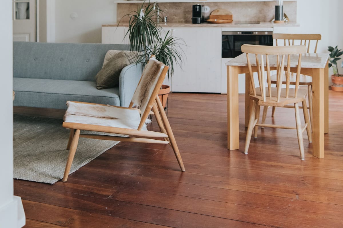 Een moderne woonkamer met een grijze bank, houten stoelen, een eettafel en een keuken op de achtergrond.