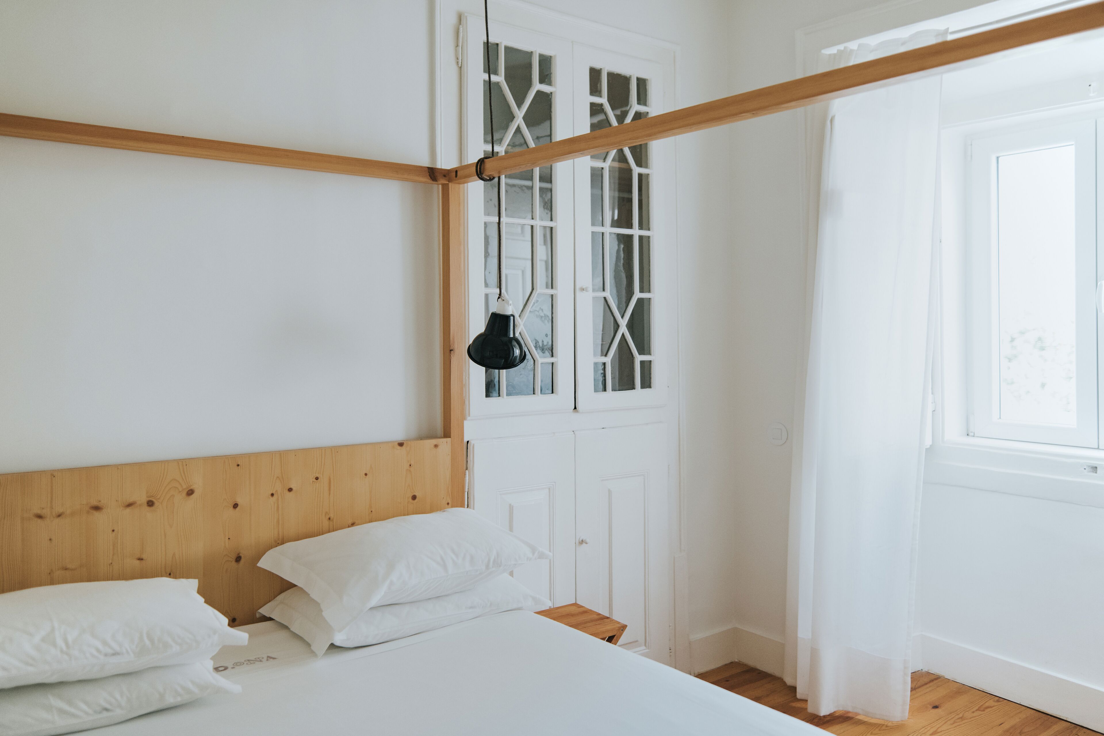Un dormitorio con cabecera de madera, ropa de cama blanca, una lámpara colgante negra y una ventana con cortinas de gasa.
